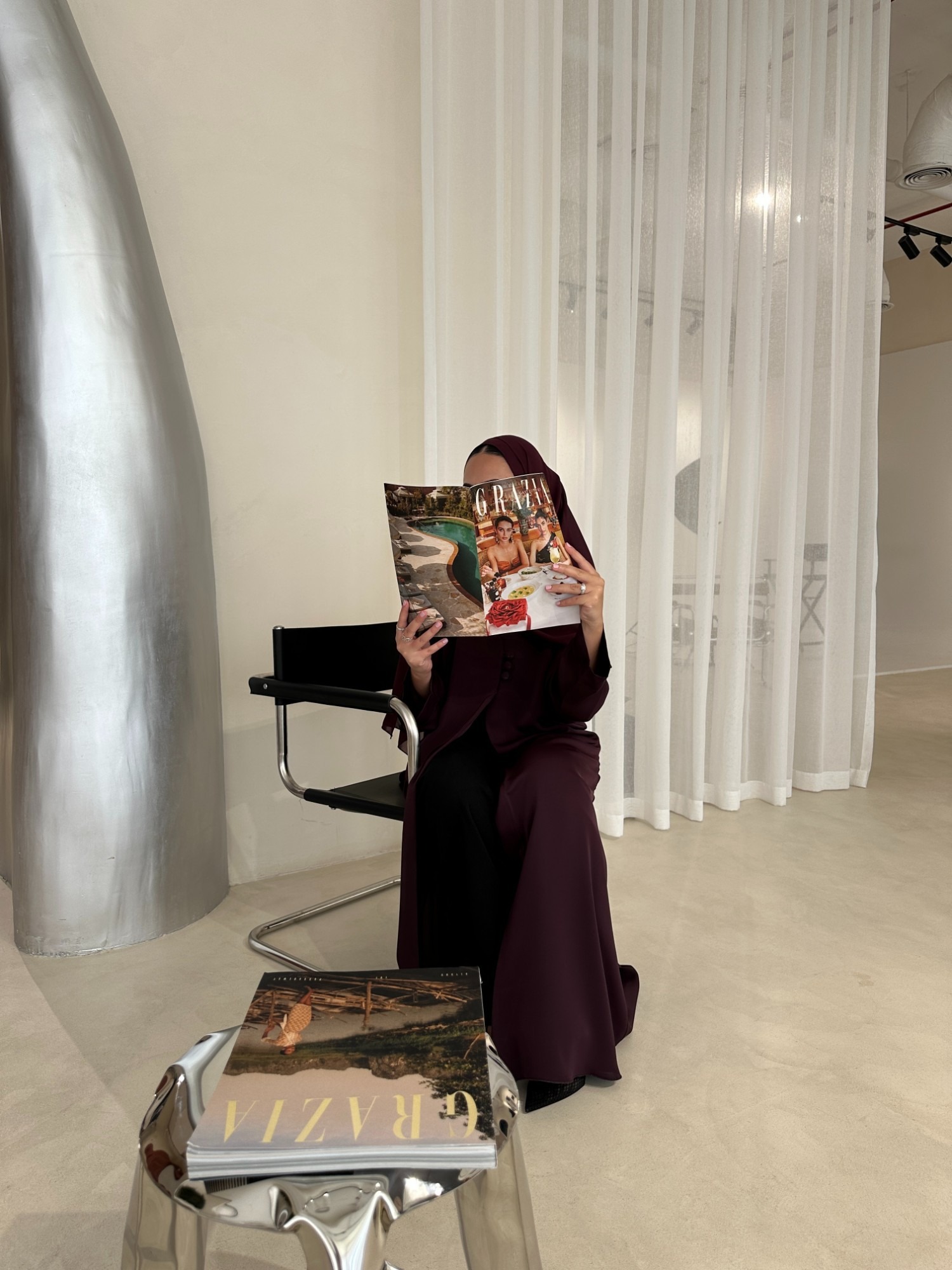 Woman in a burgundy abaya reading a magazine in a minimalist studio