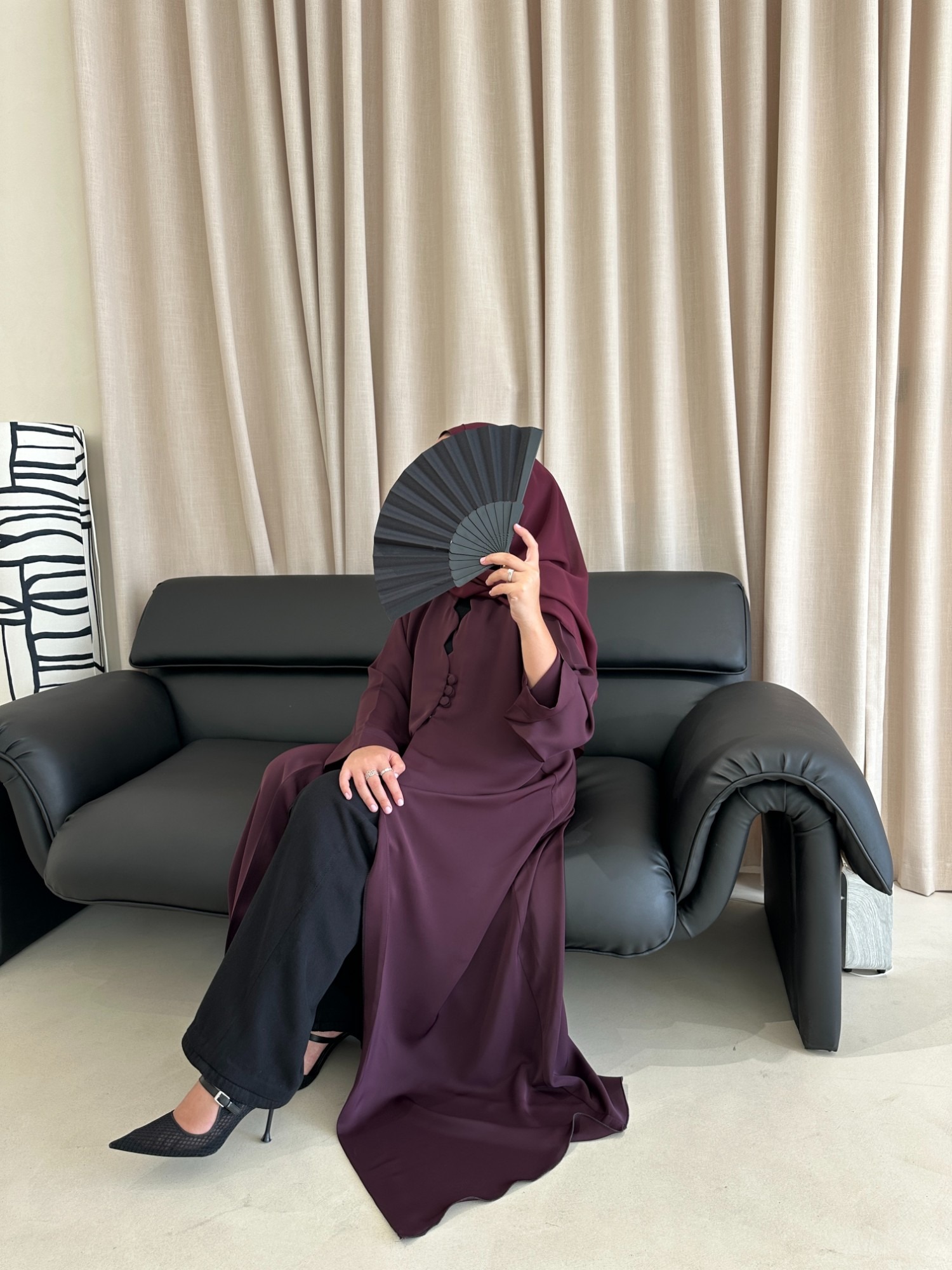 Woman in a burgundy abaya holding a black fan, seated on a black leather sofa